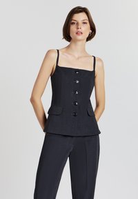 Donna con capelli corti castani che indossa un top senza maniche nero con bottoni e pantaloni neri abbinati, su uno sfondo chiaro e semplice.