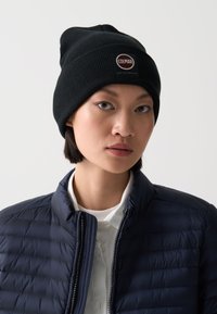 Svart stickad mössa med ribbat mönster och logopatch; tillsammans med en figursydd marinblå puffjacka och en vit skjorta under.