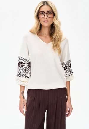 Blonde vrouw met grote schildpadkleurige bril, witte trui met zwarte patroonmouwen en bruine pijpenbroeken met krijtstrepen op een witte achtergrond.