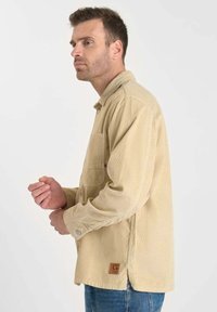 Beige corduroy jas met een knoopsluiting aan de voorkant, lange mouwen, een borstzak en een logo patch nabij de zoom. Casual pasvorm en textuurstof.