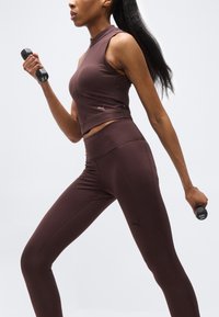 Γυναίκα με καφέ αθλητική ενδυμασία που ασκείται με αλτήρες βάρους 1 κιλού, σε μέση βήματος, μπροστά σε απλό ανοιχτό φόντο.