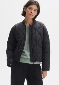 Zwarte gewatteerde bomberjack met een gestructureerde afwerking, voorzien van een ritssluiting aan de voorkant en geribde manchetten, gedragen over een lichtgroene trui.