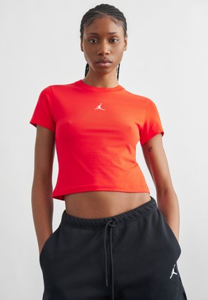 Giovane donna con capelli intrecciati che indossa una t-shirt Jordan rossa corta e pantaloni neri con coulisse, con le mani nelle tasche.