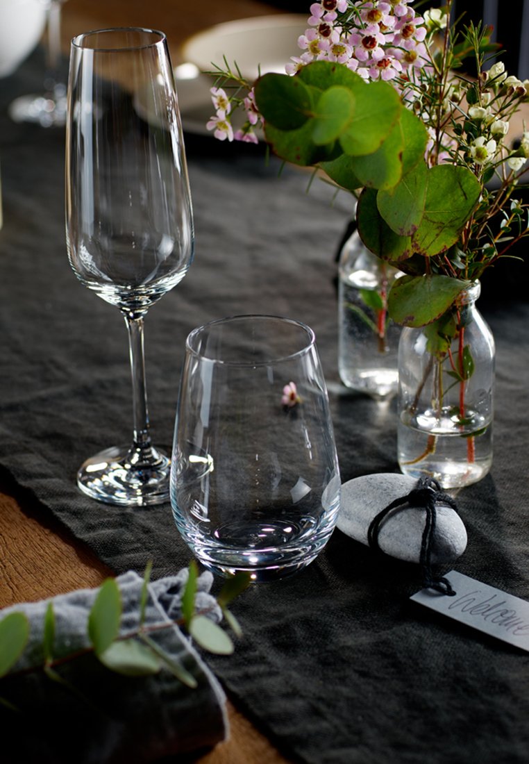 Zwei Glaswaren auf einem dunklen Tischläufer aus Leinen platziert. Ein hohes Sektglas und ein kurzer runder Tumbler, neben kleinen Blumenarrangements in Flaschen.