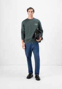 Sweatshirt verde-escuro com logotipo branco, jeans azuis e sapatos pretos. Segura uma mala preta. Design simples e casual com um ajuste relaxado.