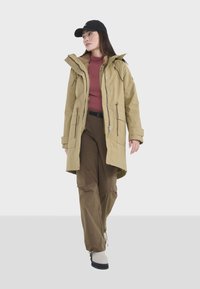 Parka longue beige avec une fermeture éclair à l'avant, deux poches latérales et une capuche, portée par-dessus un haut bordeaux et un pantalon marron, complétée par une casquette noire.
