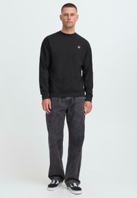 Ung mann står iført svart genser, falmede svarte jeans og svarte joggesko med hvite såler mot en enkel hvit bakgrunn.