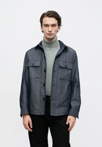 Fiatal férfi szürke garbóban és kék, patentos, zsebes farmerdzsekiben, egyszínű fehér háttér előtt áll.