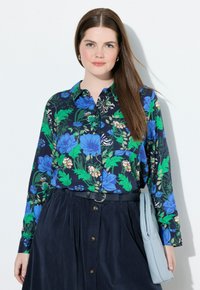 Donna con lunghi capelli castani, che indossa una camicia a bottoni floreale blu e verde, una gonna blu scuro con una cintura e tiene una clutch azzurro chiaro.