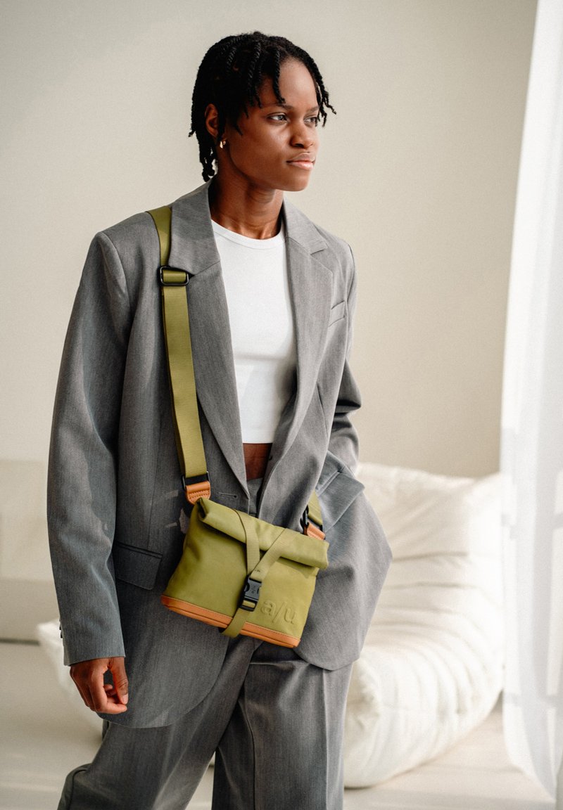 Young person wearing a gray oversized suit and white cropped top, carrying an olive green crossbody bag, standing in a bright room.