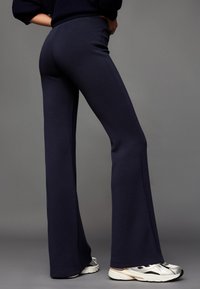 Granatowe spodnie dresowe z rozszerzanymi nogawkami wykonane z miękkiego materiału, z wysokim stanem i gładką fakturą, zestawione z białymi i srebrnymi butami sportowymi.