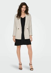 Lichtgrijze dubbelzijdige blazer met gouden knopen, scherpe revers en lange mouwen, gedragen over een zwarte plisséjurk en zwarte hakken.