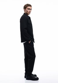 Schwarze, taillierte Shirtjacke mit einer Brusttasche, kombiniert mit lockeren schwarzen Hosen und schwarzen Schnürschuhen. Einfaches, minimalistisches Design.