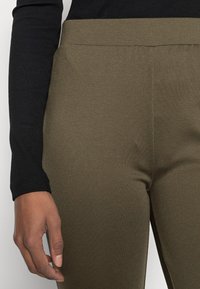 Calças verde-oliva com uma textura suave, apresentando uma cintura larga e costuras laterais. Camisola preta de manga longa com textura canelada visível perto do pulso.