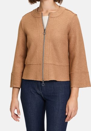 Vrouw draagt een camelkleurig jack met rits, een ronde kraag en driekwart mouwen, gecombineerd met donkerblauwe spijkerbroek.