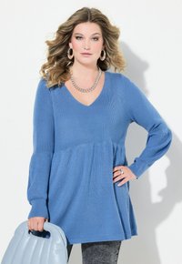 Blaue Strick-Tunika mit V-Ausschnitt, langen Ärmeln und fließendem Saum. Kombiniert mit einer hellblauen Handtasche und einer silbernen Kettenhalskette.