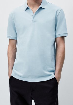 Man wearing a light blue short-sleeve polo shirt and black pants with hands in pockets, shown from neck to hips.