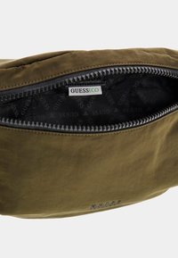 Olivgrüne Tasche mit Reißverschluss, die über ein glattes Außenmaterial und ein gemustertes schwarzes Innenfutter mit GUESS-Logo verfügt.