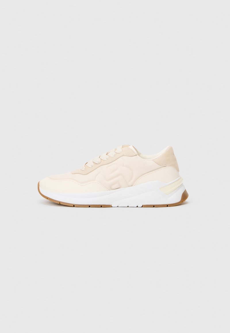 Beige sneaker fűzős elülső résszel, texturált oldalsó logóval, fehér középtalppal és világosbarna gumitalppal, szürke háttérrel.