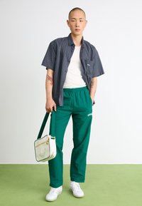 Korte mouw geruite shirt, witte tanktop, groene elastische taillebroek, witte sneakers, met een crèmekleurige tas met groene accenten en logo.