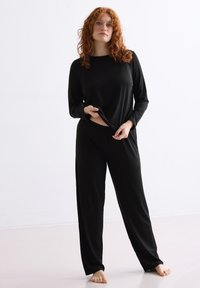 Schwarzes Loungewear-Set bestehend aus einem langärmligen Oberteil und lockeren Hosen. Weicher, glatter Stoff mit einem entspannten Schnitt und elastischem Bund.