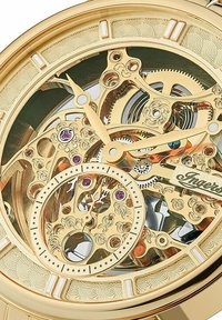 Montre mécanique en ton or avec des engrenages et des pierres précieuses complexes, visibles à travers le cadran transparent, présentant des motifs décoratifs et des marqueurs clairs.