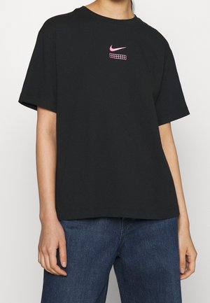 Person iført en løs sort T-shirt med korte ærmer med en lille lyserød Nike-logo og gitterdesign på øverste del af brystet, kombineret med blå jeans.