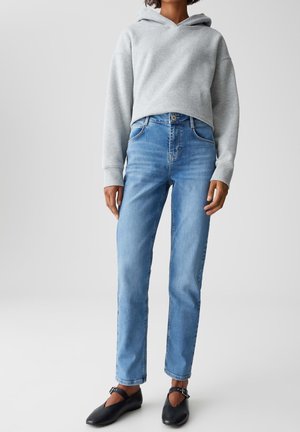 Persoon in lichtblauwe high-waisted jeans, een grijze cropped hoodie en zwarte platte schoenen met enkeldraagriemen, staande tegen een effen achtergrond.