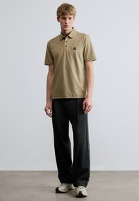 Béžové bavlnené polo tričko s trojguľovým zapínaním, čierna logová nášivka na ľavej strane hrudníka, nosené s voľnými čiernymi nohavicami a bielymi teniskami.