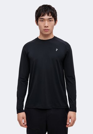Jeune homme debout, face à nous, portant un maillot de sport noir à manches longues avec un petit logo sur la poitrine.
