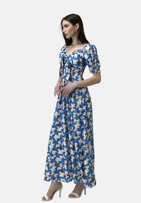 Maxikleid in Weiß mit blau-braunem Blumenmuster, kurzen Puffärmeln und tailliertem Oberteil mit Bindeband. Kombiniert mit weißen High Heels.