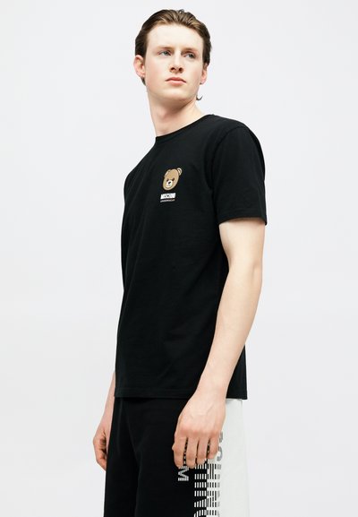 Νεαρός άνδρας που φοράει μαύρο t-shirt Moschino με λογότυπο αρκουδάκι και ασπρόμαυρο σορτς μπροστά σε απλό φωτεινό φόντο.