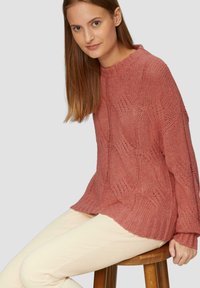 Teksturowany sweter w kolorze różowym, wykonany na drutach, o luźnym kroju, szerokich rękawach i prążkowanym dekolcie, zestawiony ze kremowymi spodniami.