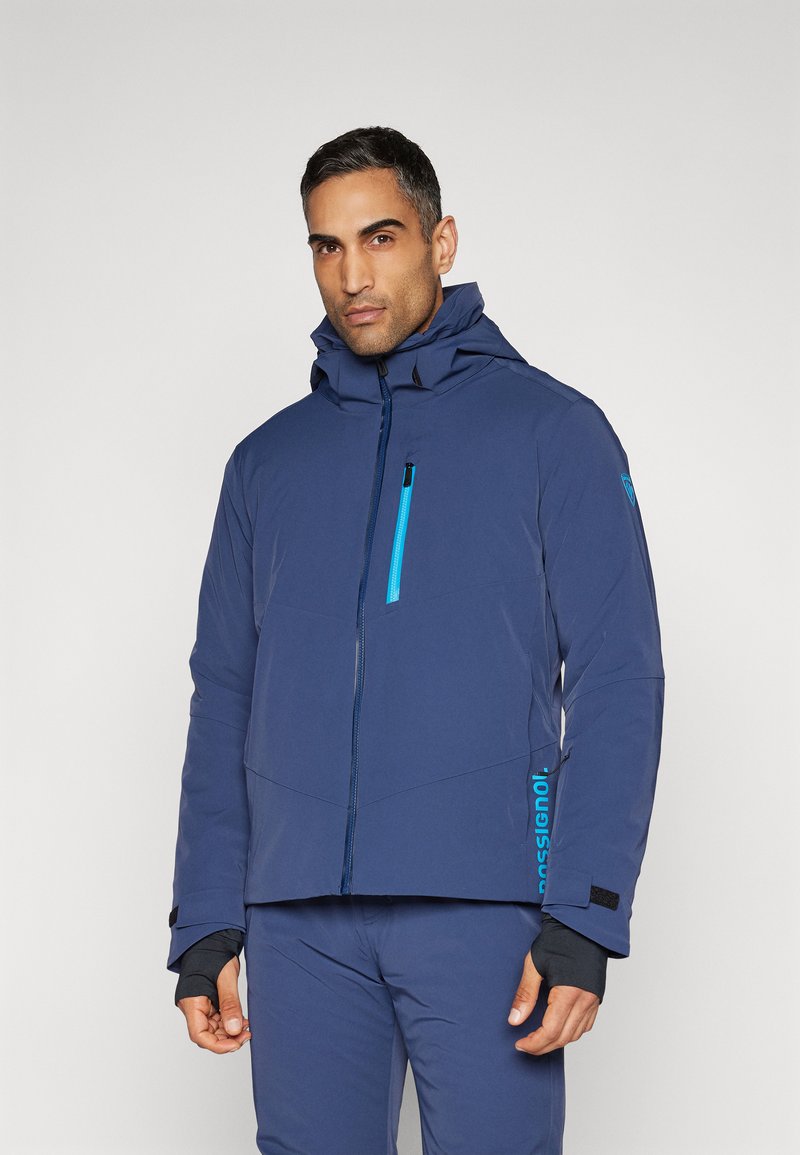 Veste isolante bleu marine avec capuche, présentant une fermeture éclair bleue contrastante, des poches et un logo sur le côté. Tissu texturé avec des poignets.