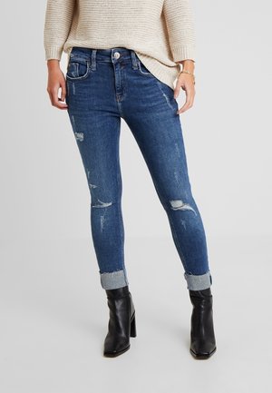 Vaqueros ajustados azul con detalles desgastados, dobladillo enrollado y diseño de cinco bolsillos. Llevados con botas negras hasta el tobillo y un suéter de punto beige.