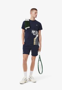 Hombre en un conjunto de tenis azul marino sosteniendo una raqueta de tenis verde, con una toalla sobre el hombro y zapatillas blancas, contra un fondo blanco.