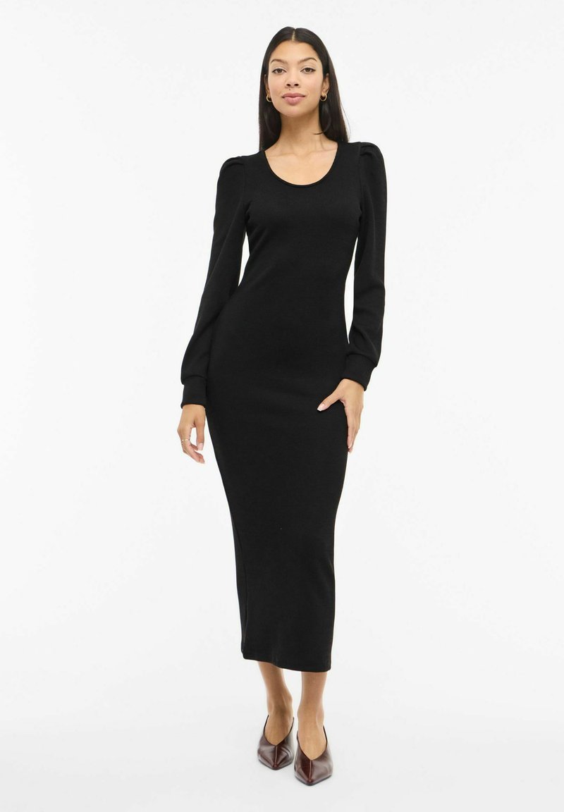 Musta pitkähihaista neulemekkoa, jossa on pyöreä pääntie, vartalonmyötäinen siluetti ja pussihihat, yhdistetty viininpunaisiin kärjistettyihin korkeakorkoisiin kenkiin.