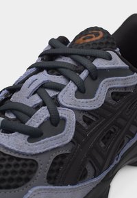 Primer plano de zapatilla deportiva negra y gris con cordones negros y parte superior de malla texturizada, mostrando detalles de costuras y logo en la lengüeta.