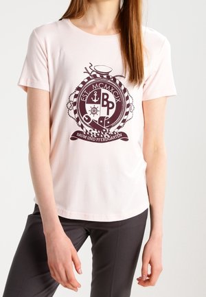 Kobieta nosząca jasnoróżowy t-shirt z krótkim rękawem z ciemnym emblematem żeglarskim i napisem "BAUM UND PFERDGARTEN" z przodu, w połączeniu z ciemnymi spodniami.