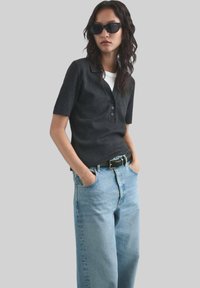 Zwarte polo met korte mouwen en drie knopen, gecombineerd met lichtblauwe rechte jeans en een zwarte riem, gedragen met donkere zonnebril.