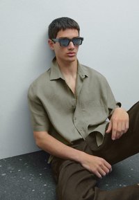La camicia khaki a maniche corte presenta una tasca ed è abbinata a pantaloni marrone scuro. Il modello indossa occhiali da sole scuri a forma quadrata, seduto contro un muro chiaro.