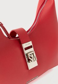 Κόκκινη δερμάτινη τσάντα με υφή, ασημένιο φερμουάρ, εξέχον λουκέτο με το χρυσό λογότυπο SM και πλακέτα με το όνομα της μάρκας Steve Madden.