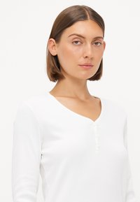 Vit långärmad topp med V-ringning och placket med fyra knappar. Tillverkad av ribbat tyg, med en mjuk textur och en åtsittande siluett.