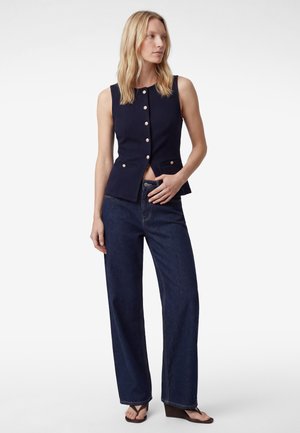 Donna bionda che indossa un gilet senza maniche blu navy con bottoni, jeans scuri a gamba larga con vita alta e sandali con zeppa marroni, in piedi con una mano vicino alla vita.