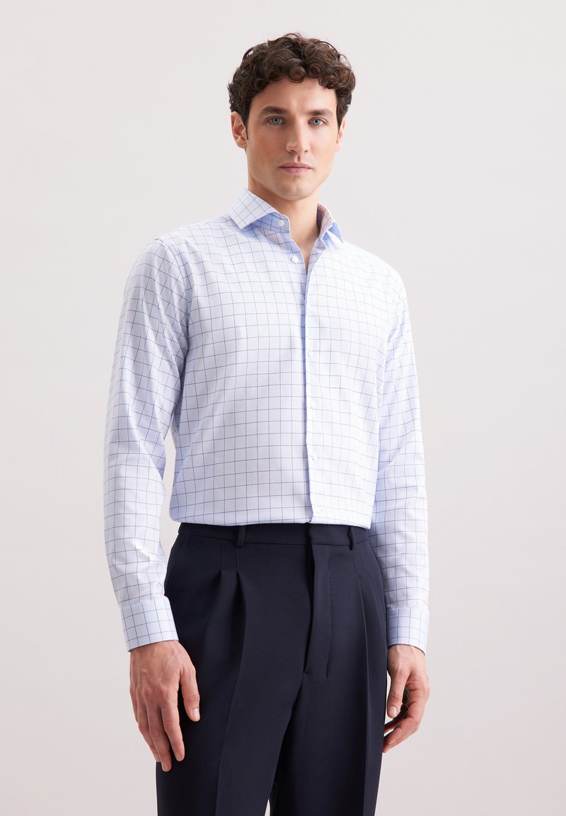 Camicia bianca con motivo a griglia blu, maniche lunghe, colletto abbottonato; abbinata a pantaloni blu navy, con pences e tessuto liscio.