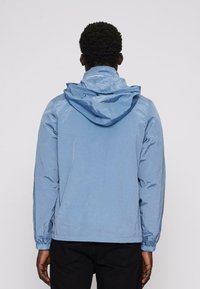 Svetle modrá bunda s kapucí s lesklou texturou, elastickými manžetami a spodním lemem. Má minimalistický design, vhodný pro volnočasové nošení.