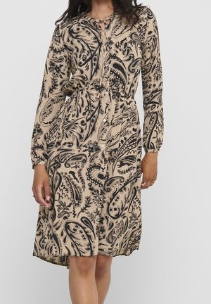 Beige jurk tot op de knie met zwart paisley-patroon, lange mouwen, knoopsluiting aan de voorkant en een getailleerde taille, gedragen door een staande persoon.