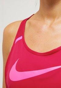 Rode sportbeha gemaakt van rekbaar materiaal, met een contrasterend roze Nike-logo, ronde halslijn en brede bandjes. Gladde textuur, atletisch ontwerp.
