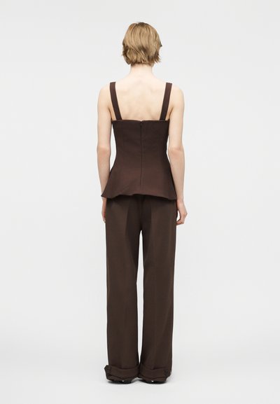 Άτομο με κοντά ανοιχτόχρωμα μαλλιά στέκεται γυρισμένο με την πλάτη, φορώντας μπεζ αμάνικο τοπ με φερμουάρ και παντελόνι φαρδύ με μανσέτες στο κάτω μέρος.