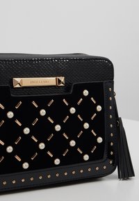 Czarna torebka na ramię z teksturowanym wykończeniem, złotymi akcesoriami, sztucznymi perłami i dekoracyjnymi ćwiekami. Uzupełniona frędzlami z boku.